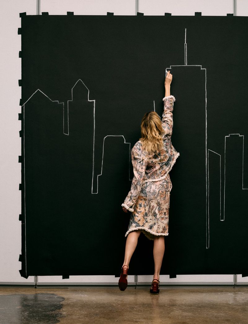Angèle viewed from behind reaching up to draw a white chalk skyscraper outline on a large black studio backdrop.