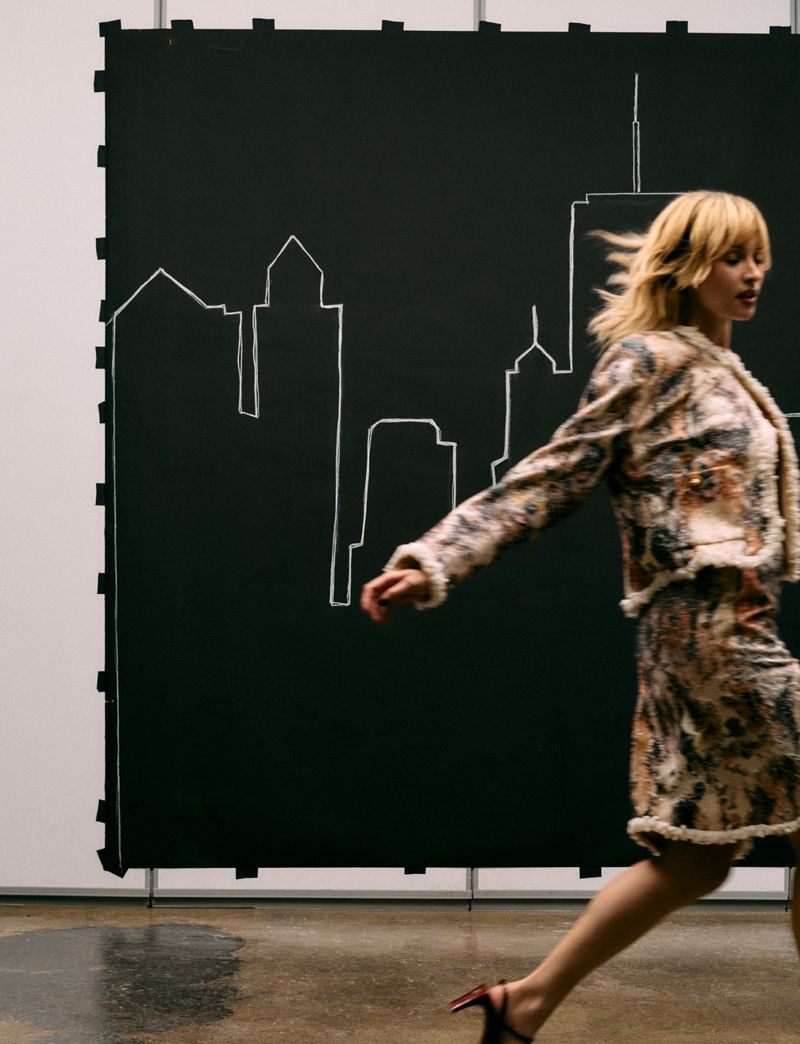 Angèle walking across a studio in a patterned textured skirt suit before a black backdrop with an illustrated city skyline.