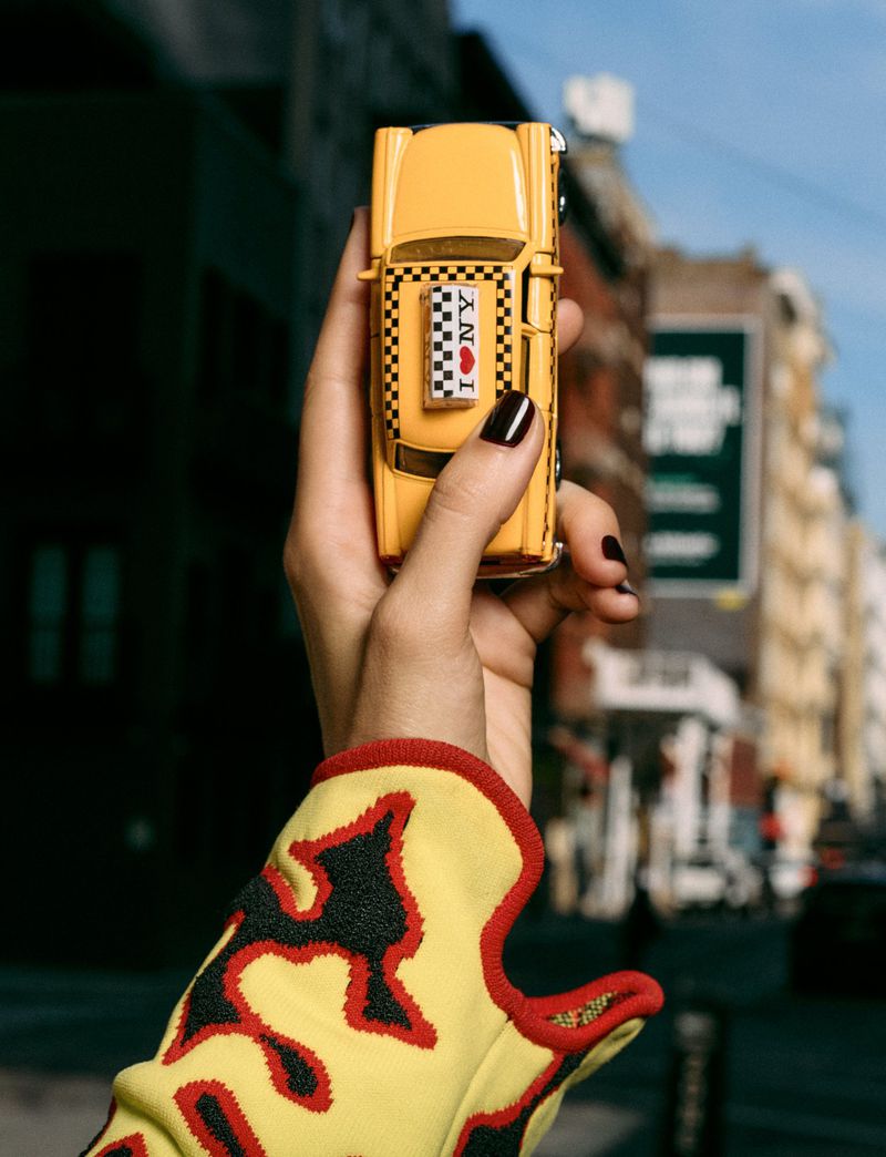 Hand with dark manicure holding a miniature yellow New York taxi toy against an out-of-focus city backdrop.