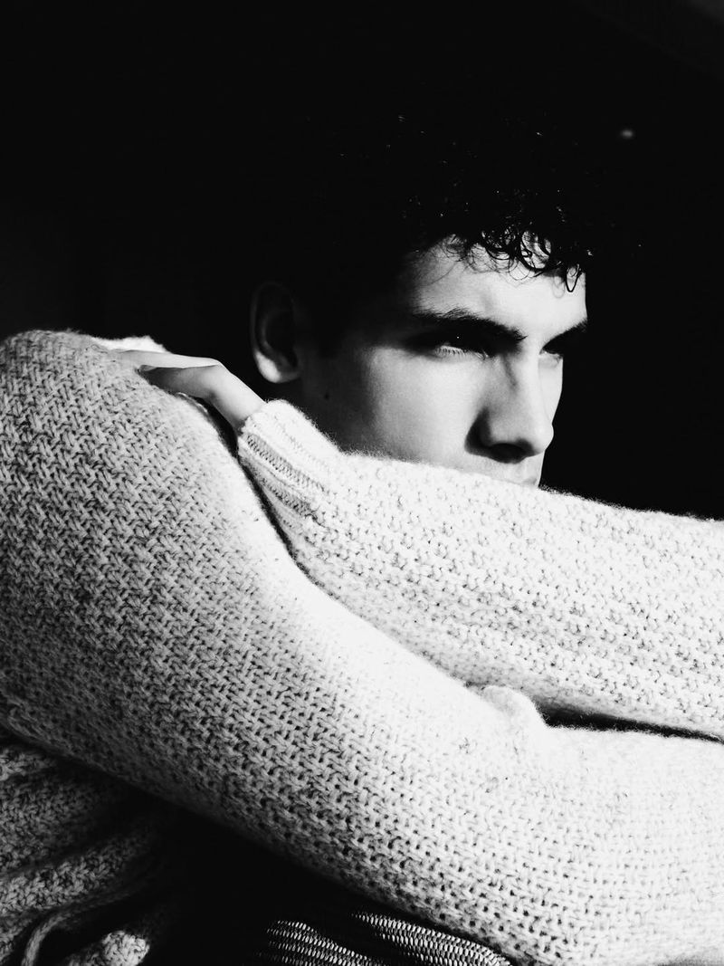 Black and white portrait of a young man wearing a heavy knit sweater with arms crossed against a dark background.