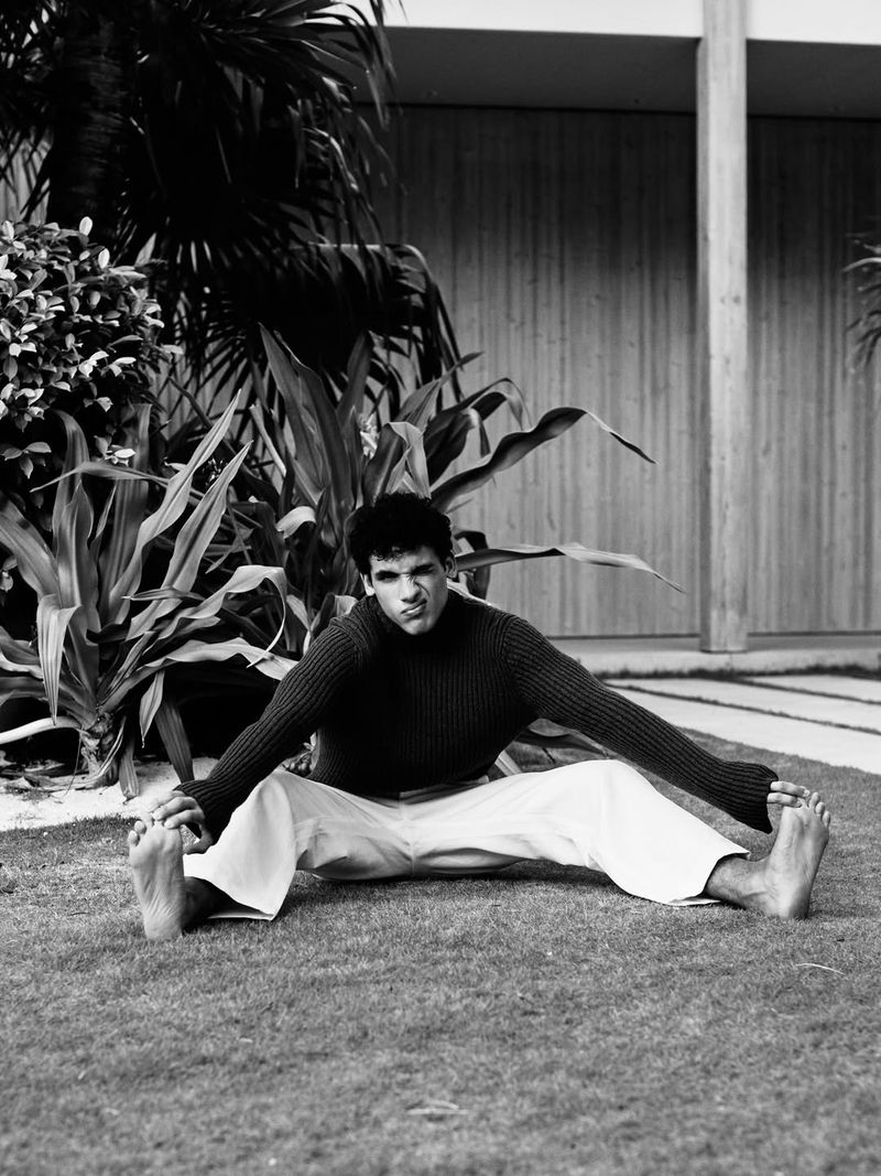 Black and white portrait of a barefoot man sitting on a lawn in a wide straddle stretch holding his feet.