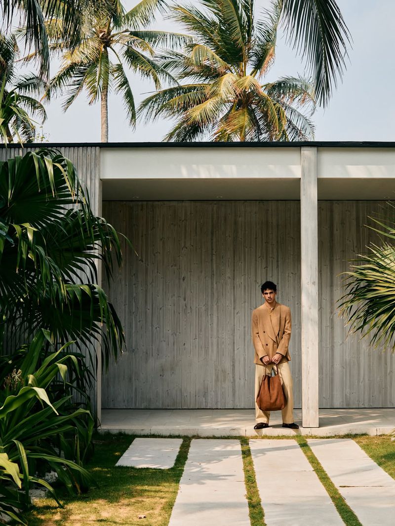 Model standing outside a modern pavilion in a camel blazer and wide trousers, holding a brown tote bag beside palm trees.