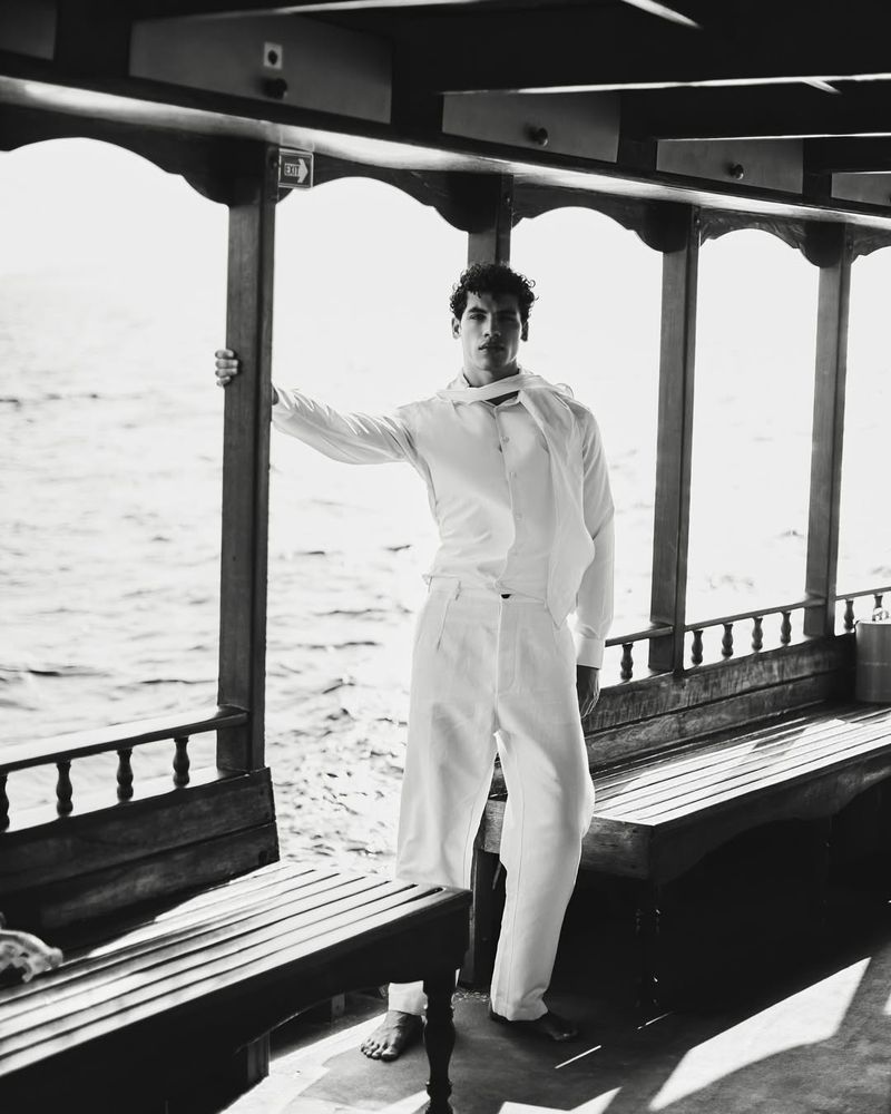Monochrome portrait of a man standing barefoot on a boat deck wearing an all-white shirt, scarf, and wide-leg trousers.