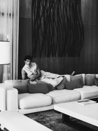A young man reclining on a large white sectional sofa in an upscale living room with dark vertical wall art in black and white.