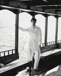 Monochrome portrait of a man standing barefoot on a boat deck wearing an all-white shirt, scarf, and wide-leg trousers.