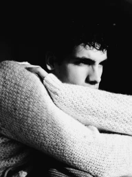 Black and white portrait of a young man wearing a heavy knit sweater with arms crossed against a dark background.