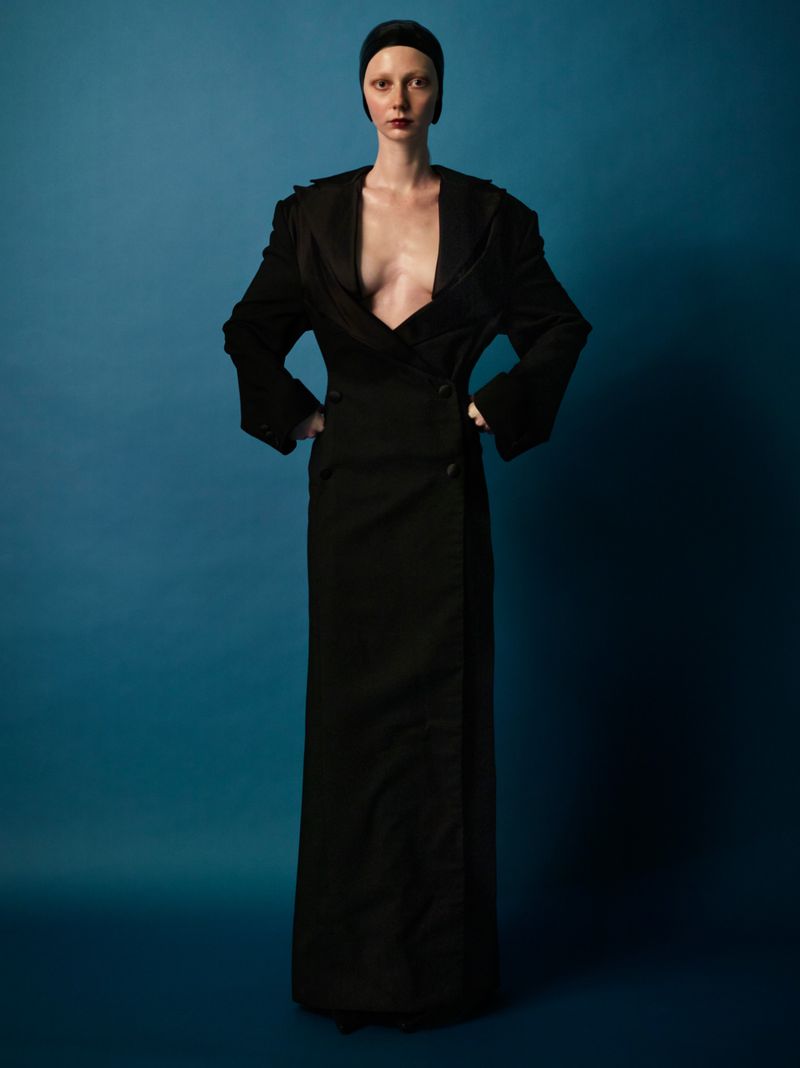 Model wearing a floor-length black double-breasted coat dress and black skullcap posing against a rich blue studio backdrop.