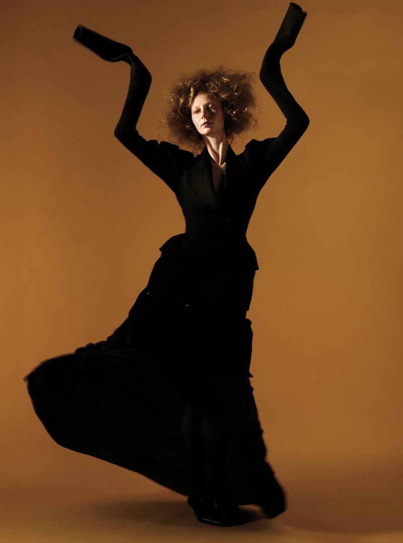 Model posing with raised arms wearing a tailored black ensemble with elongated sleeves against an amber backdrop.
