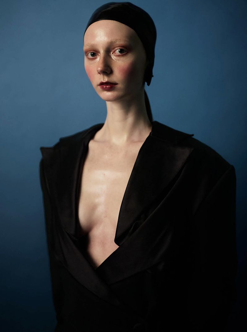 Model wearing a black open-front tailored jacket and sleek black headwrap with high-gloss editorial makeup against a blue backdrop.