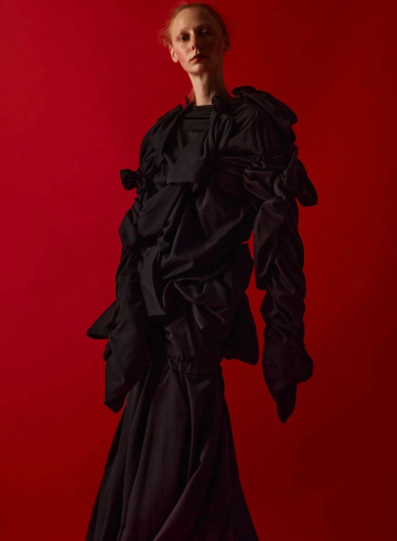 Model wearing an avant-garde black ruffled sculptural gown posing against a solid red studio backdrop.
