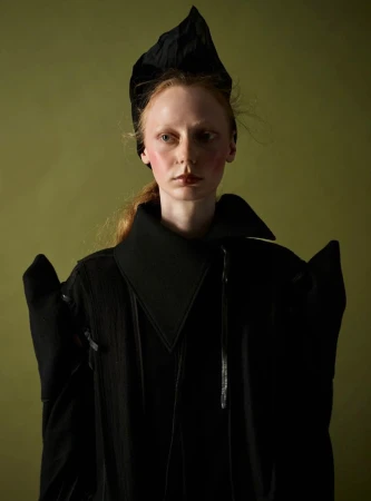Woman with dramatic blush and a sculptural black headpiece wearing a structured black coat against an olive green backdrop.