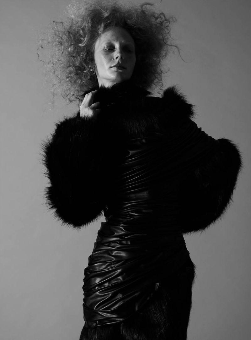 Model with voluminous curly hair wearing a black fur jacket and ruched black dress against a grey studio backdrop.