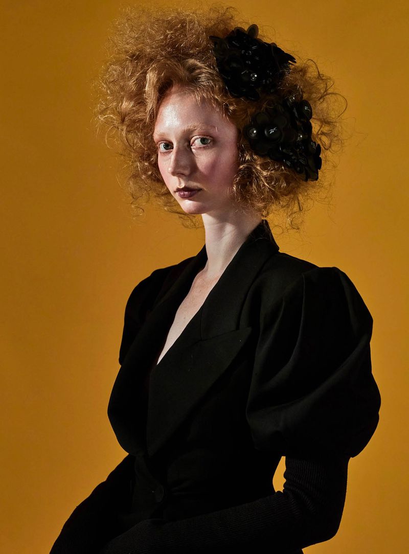 A red-haired woman wearing a black puff-sleeve jacket and a dark floral headpiece against a mustard yellow studio backdrop.