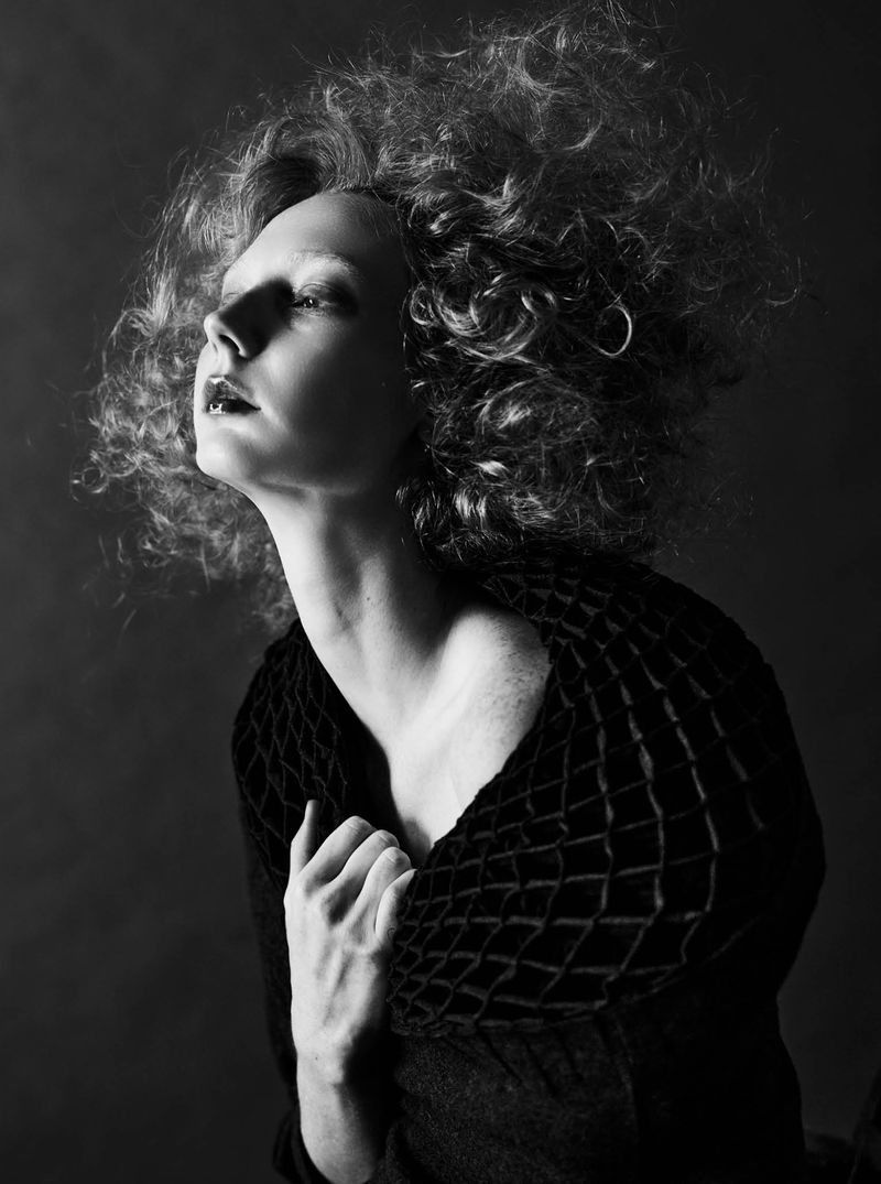 Black and white portrait of a woman with voluminous curly hair looking upward in dramatic low-key light, wearing a textured dark cape.