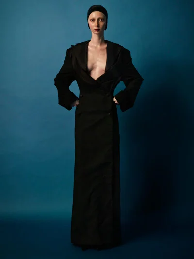 Model wearing a floor-length black double-breasted coat dress and black skullcap posing against a rich blue studio backdrop.