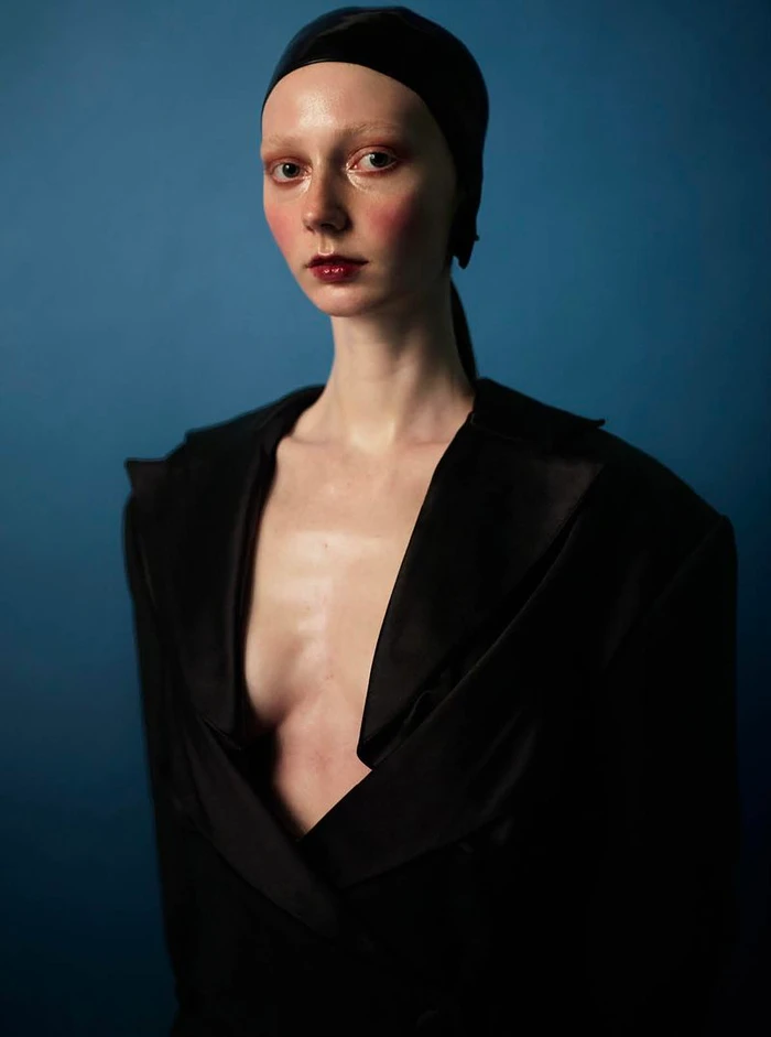 Model wearing a black open-front tailored jacket and sleek black headwrap with high-gloss editorial makeup against a blue backdrop.