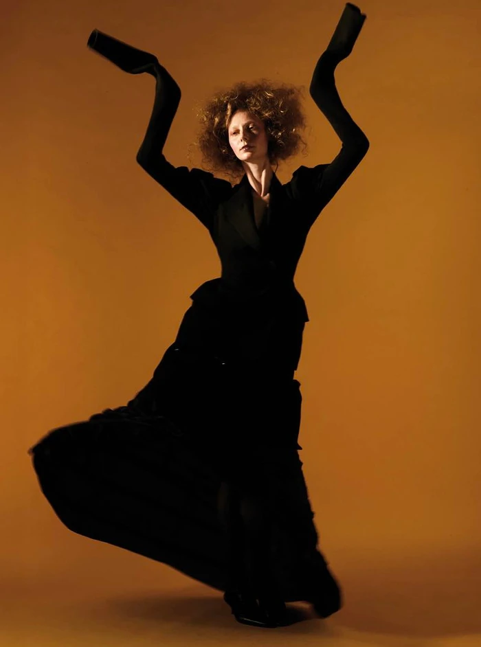 Model posing with raised arms wearing a tailored black ensemble with elongated sleeves against an amber backdrop.
