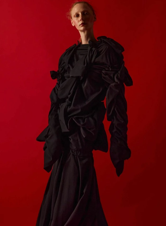 Model wearing an avant-garde black ruffled sculptural gown posing against a solid red studio backdrop.