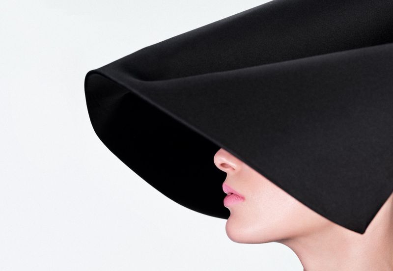 Profile close-up of a woman wearing a sculptural oversized black hat covering her eyes against a solid white studio background.