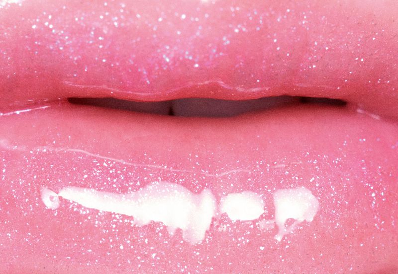 Extreme close-up of glossy pink lips with fine glitter and a white cosmetic cream streak along the lower lip.