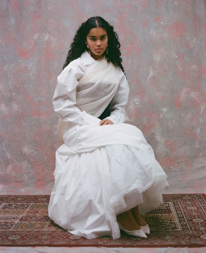 Model Ingrid Fernandes in a white layered dress seated against a pink and grey mottled backdrop for Ladygunn Magazine.