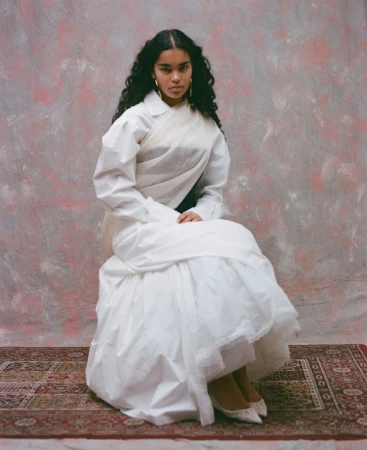 Model Ingrid Fernandes in a white layered dress seated against a pink and grey mottled backdrop for Ladygunn Magazine.