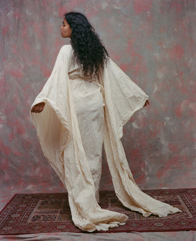 Model Ingrid Fernandes seen from behind in a flowing off-white gown with floor-length wing sleeves on an ornate patterned rug.