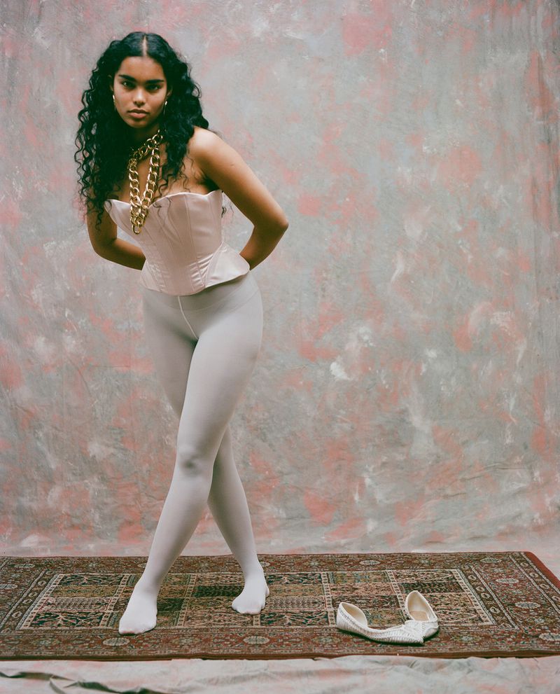 Model Ingrid Fernandes posing in a pale satin corset and white tights on an ornate rug in front of a textured backdrop.