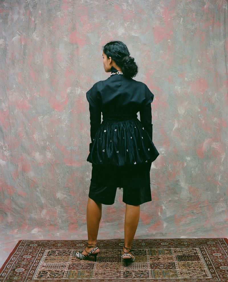 Ingrid Fernandes seen from behind wearing an embellished black peplum top and shorts standing on an ornate rug.