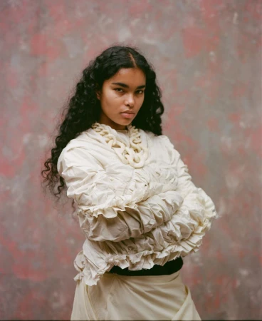 Portrait of a young woman with dark curly hair wearing a light colored, ruffled top and skirt against a mottled pink and grey background.