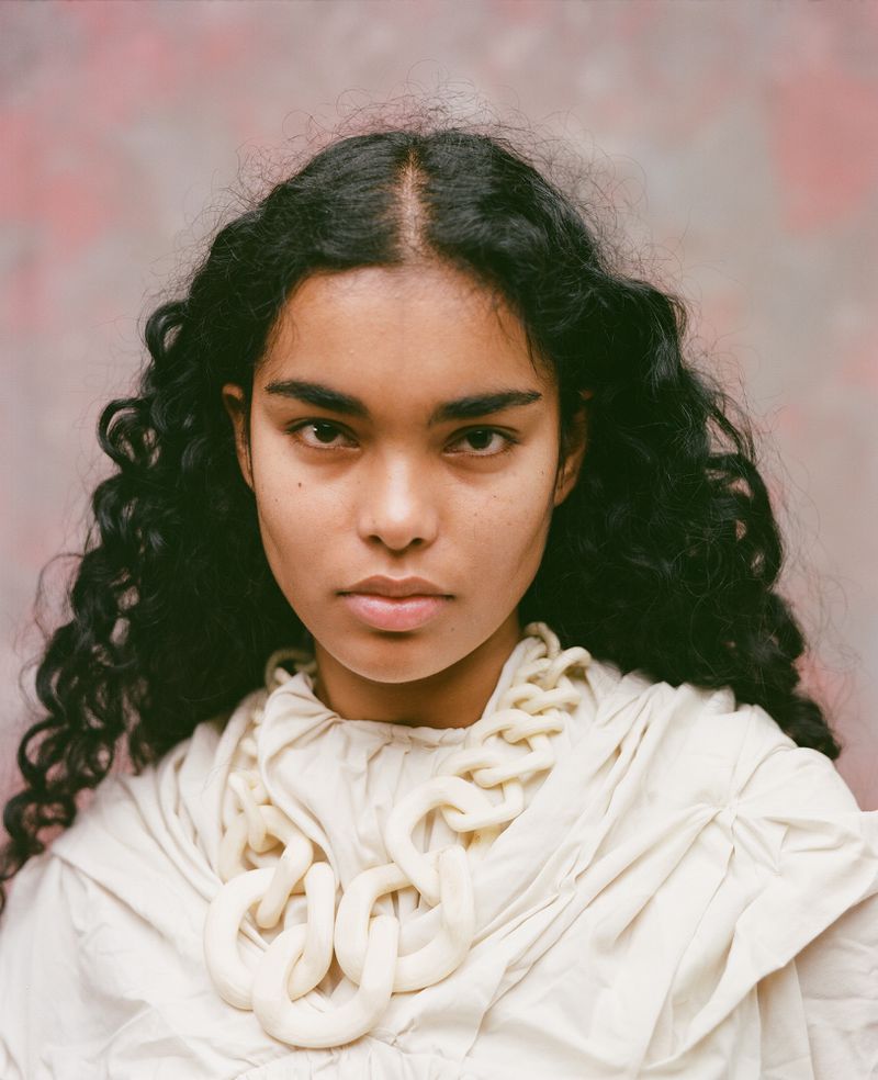 Model Ingrid Fernandes with long dark curly hair wearing an off-white draped top and a large matching chunky chain necklace.