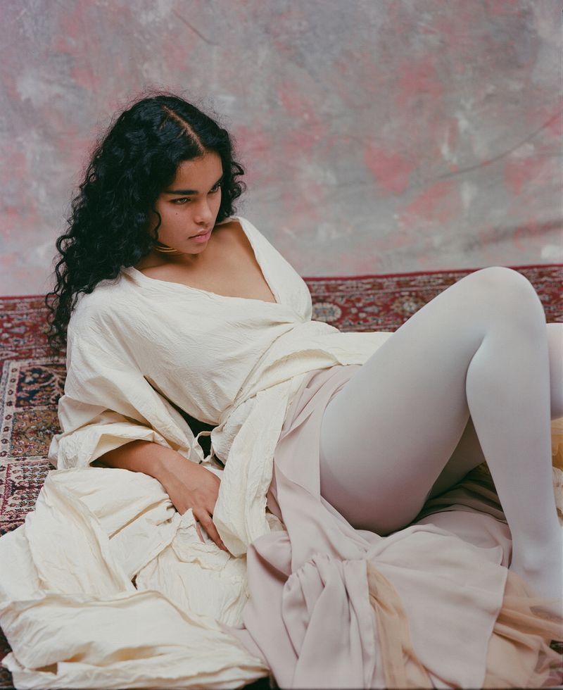 Woman with dark curly hair poses in white robe and tights on ornate carpet in a photo studio. Light and airy, fashion portrait.