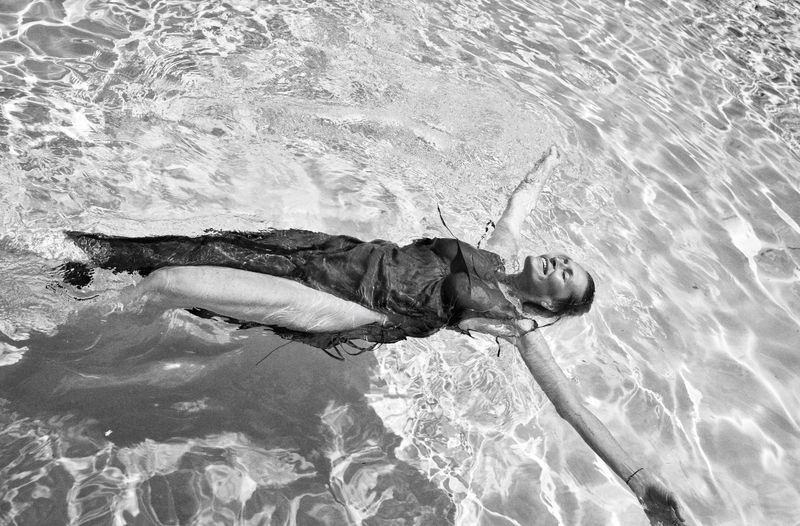 Estelle Lefébure smiling while floating on her back in a sunlit swimming pool wearing a dark sleeveless dress.