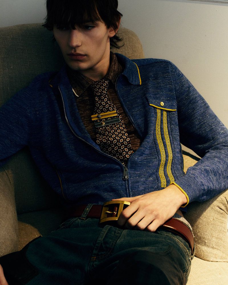 Young man seated in a fabric armchair wearing a blue zip knit sweater with yellow trim, patterned necktie, and brown leather belt.
