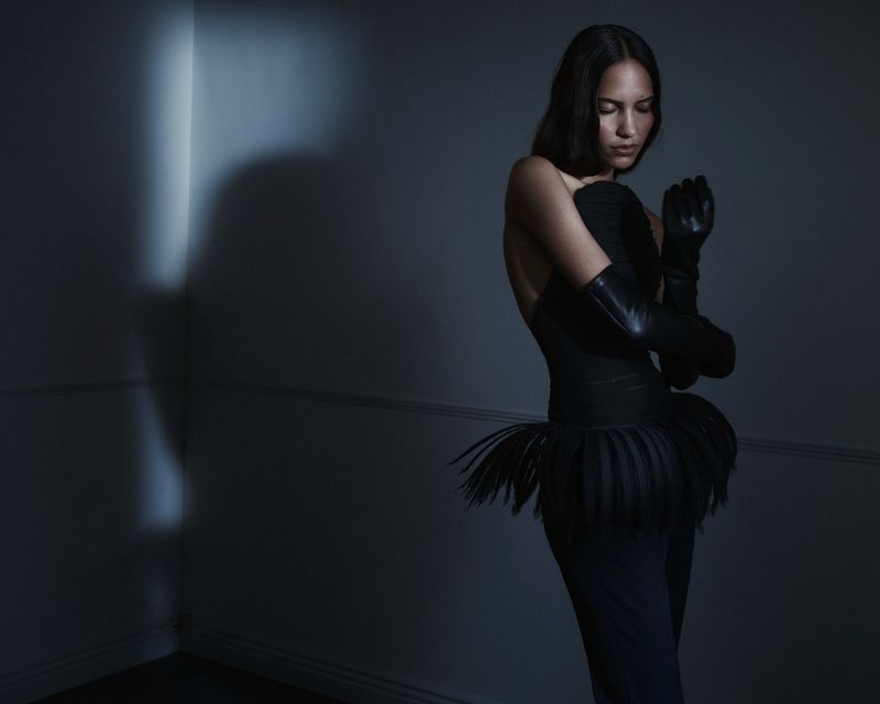 Model wearing a black strapless outfit with a fringe peplum and long black leather gloves in a dimly lit blue-toned room.