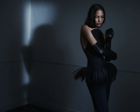 Model wearing a black strapless outfit with a fringe peplum and long black leather gloves in a dimly lit blue-toned room.