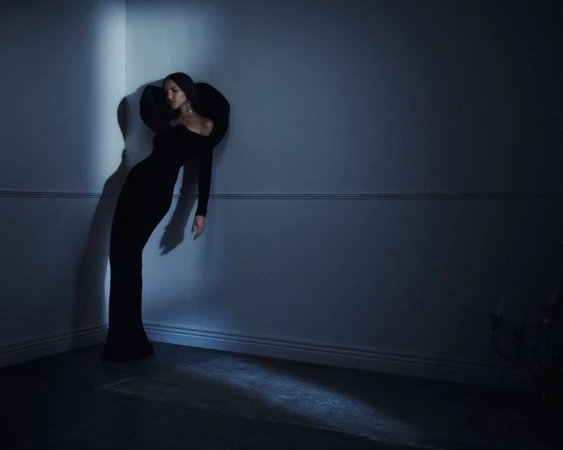 Woman in a dramatic black sculptural evening gown leaning against a wall corner in a dim, moody interior room.