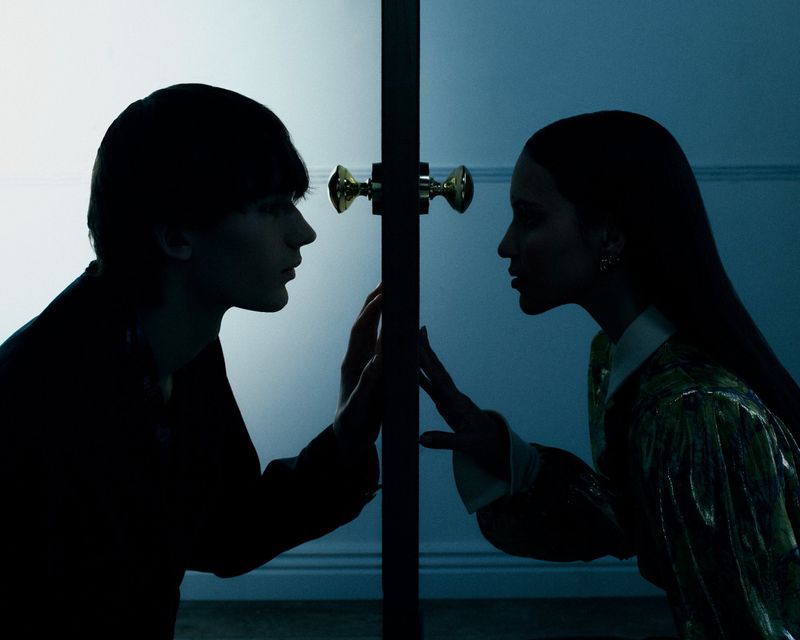 Profile silhouettes of a young man and woman placing their hands against opposite sides of a doorway under cool blue light.