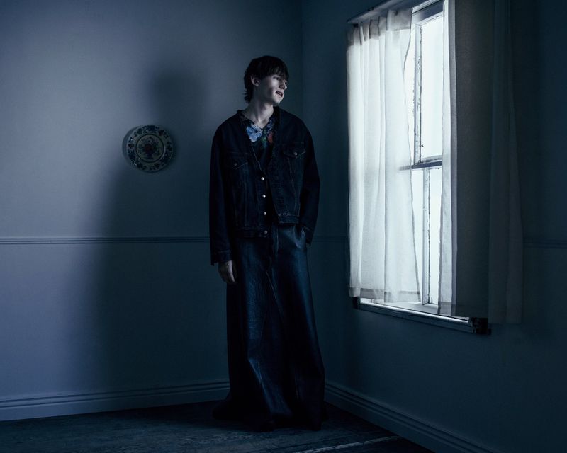 Model Noah Bates standing beside a curtained window in a dark jacket and floor-length dark skirt in a moody blue interior.