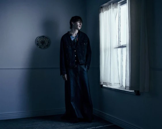 Model Noah Bates standing beside a curtained window in a dark jacket and floor-length dark skirt in a moody blue interior.