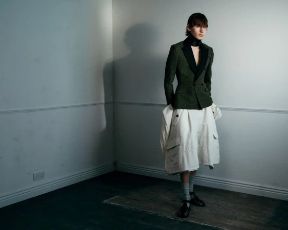Model standing in a room corner wearing a dark green tailored blazer, black neckband, wide white shorts, and black cage sandals.