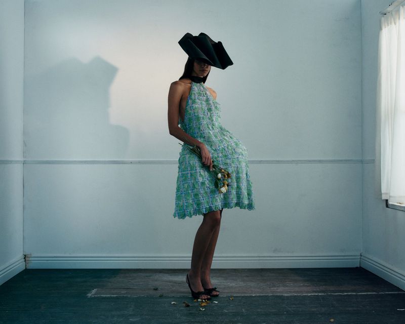 Model in a patterned green halter dress and black sculptural hat holding small flowers indoors