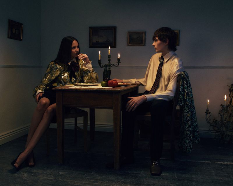 Man and woman seated at a wooden dining table lit by candles in a moody dark room for a fashion editorial.