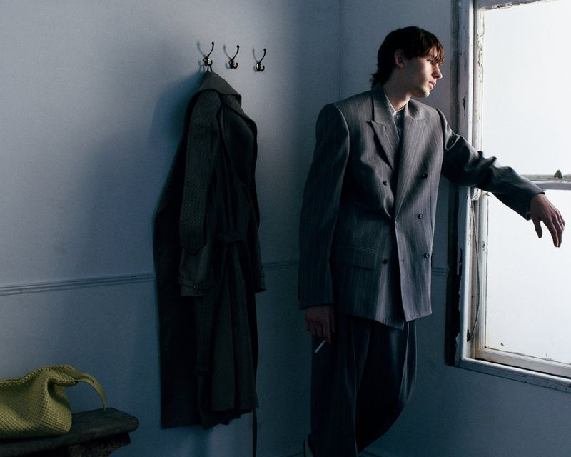 Man wearing an oversized grey double-breasted suit standing beside an open window with a hanging coat behind him