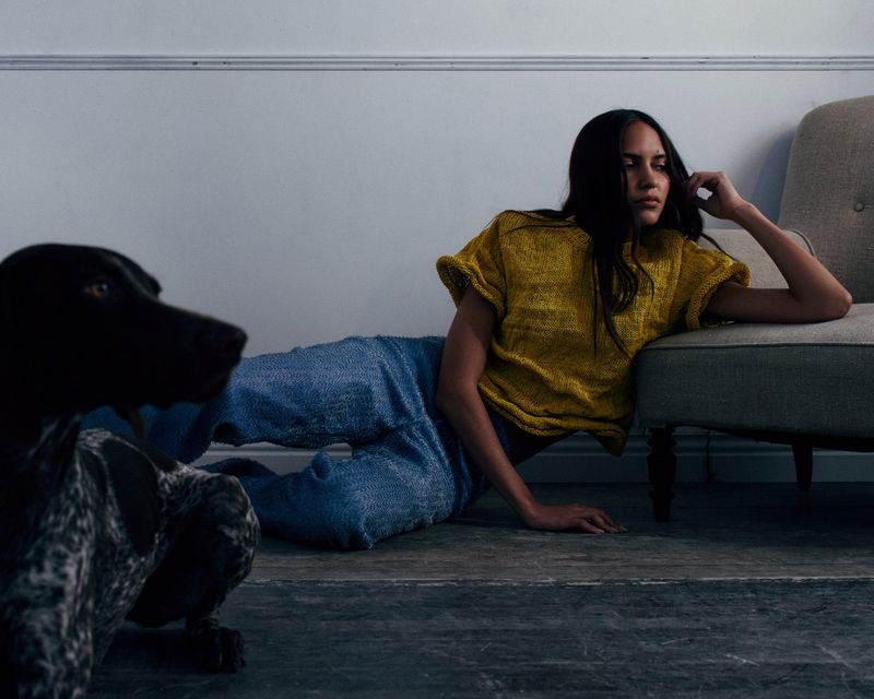 A woman in a yellow textured top and blue jeans reclines on a wooden floor beside an armchair and a speckled dog.