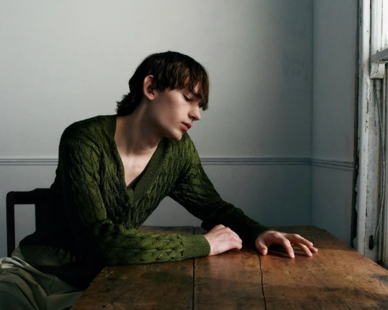 A young man with dark hair wearing an olive green cable-knit sweater leans over a rustic wooden table indoors.
