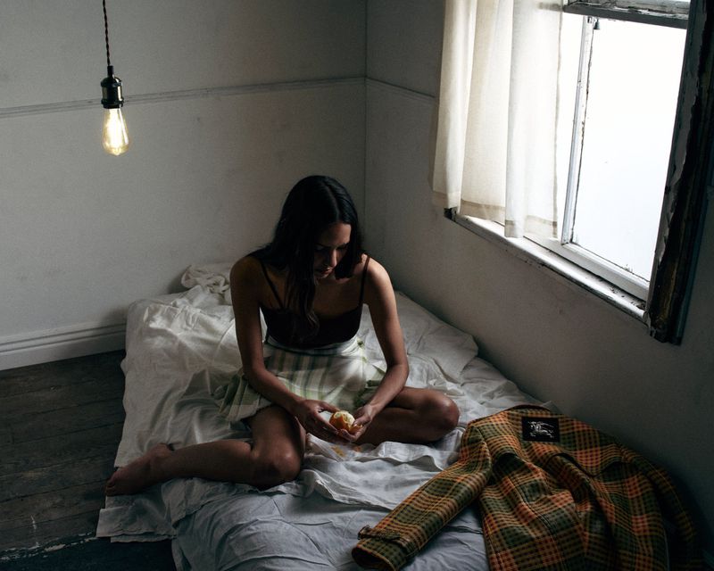 Woman sitting barefoot on a bed holding fruit near a plaid coat under a hanging lightbulb in a bedroom.