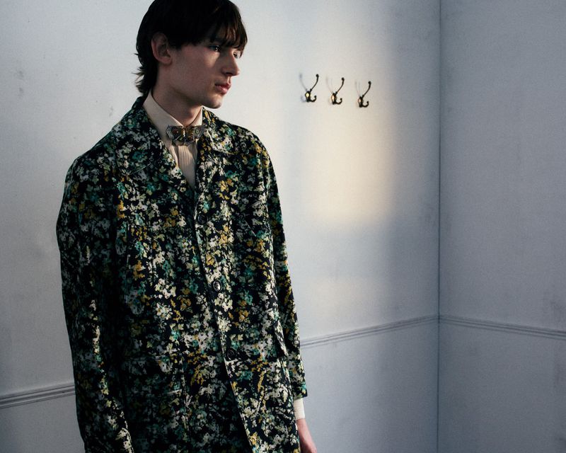 A young man in a dark floral patterned coat and collared shirt stands in profile beside three wall hooks against a white wall.