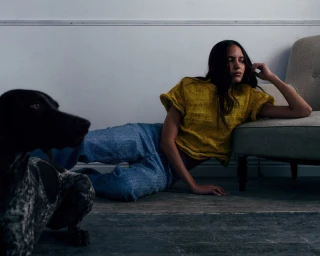 A woman in a yellow textured top and blue jeans reclines on a wooden floor beside an armchair and a speckled dog.