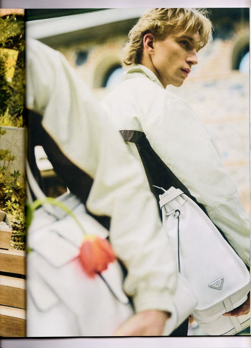 Model Aleksander Lukowski wearing an off-white jacket and carrying a white shoulder bag with an orange tulip in foreground.
